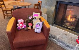 Stuffed animals sitting in a chair together by a fireplace.